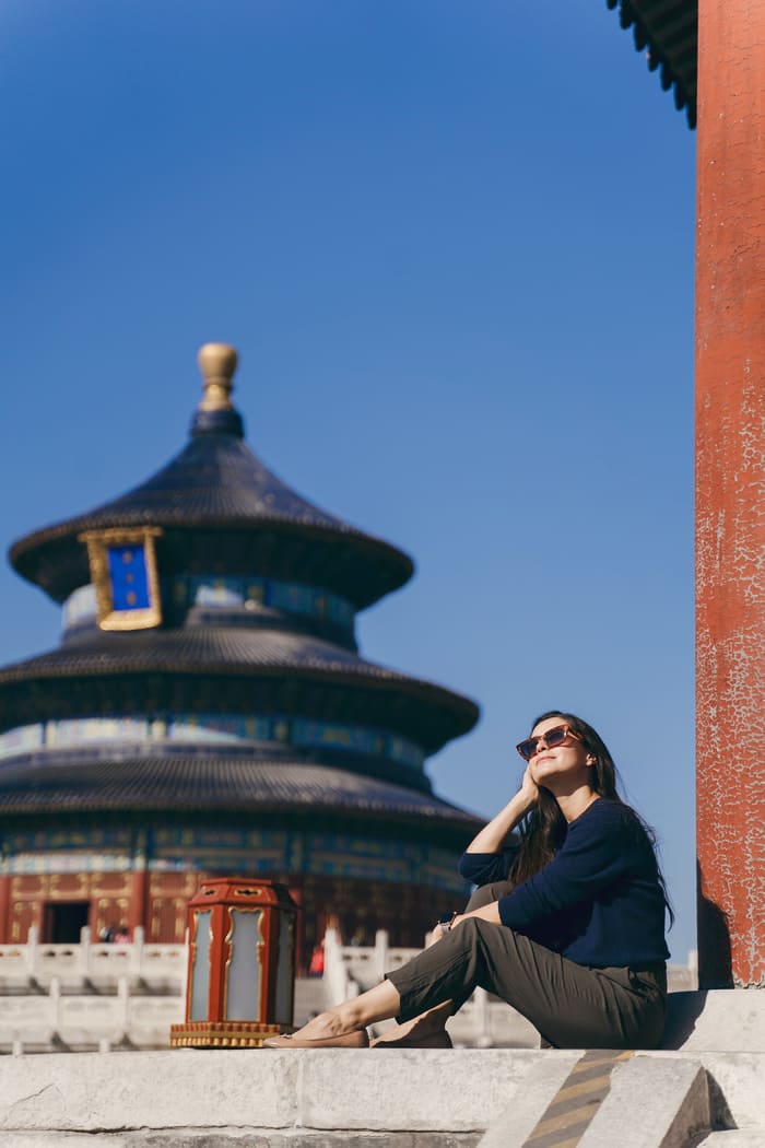 Mujer turista observa sentada hacia arriba y detras un templo en colores oscuros y rojos en la base.