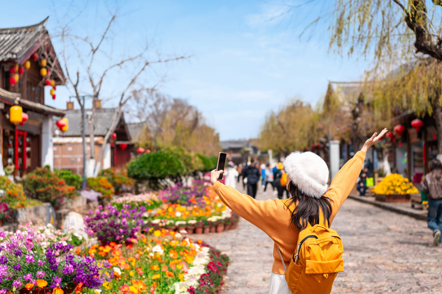 Fotografía de una mujer con sueter y mochila amarillos, frente a un templo rodeado de flores.